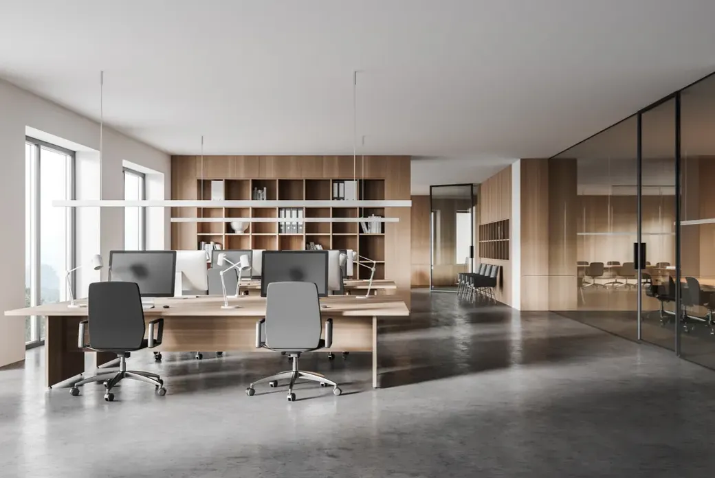 Modern office with wooden shelves, large windows, and glass partitions. Features multiple desks with computers and swivel chairs on a concrete floor.