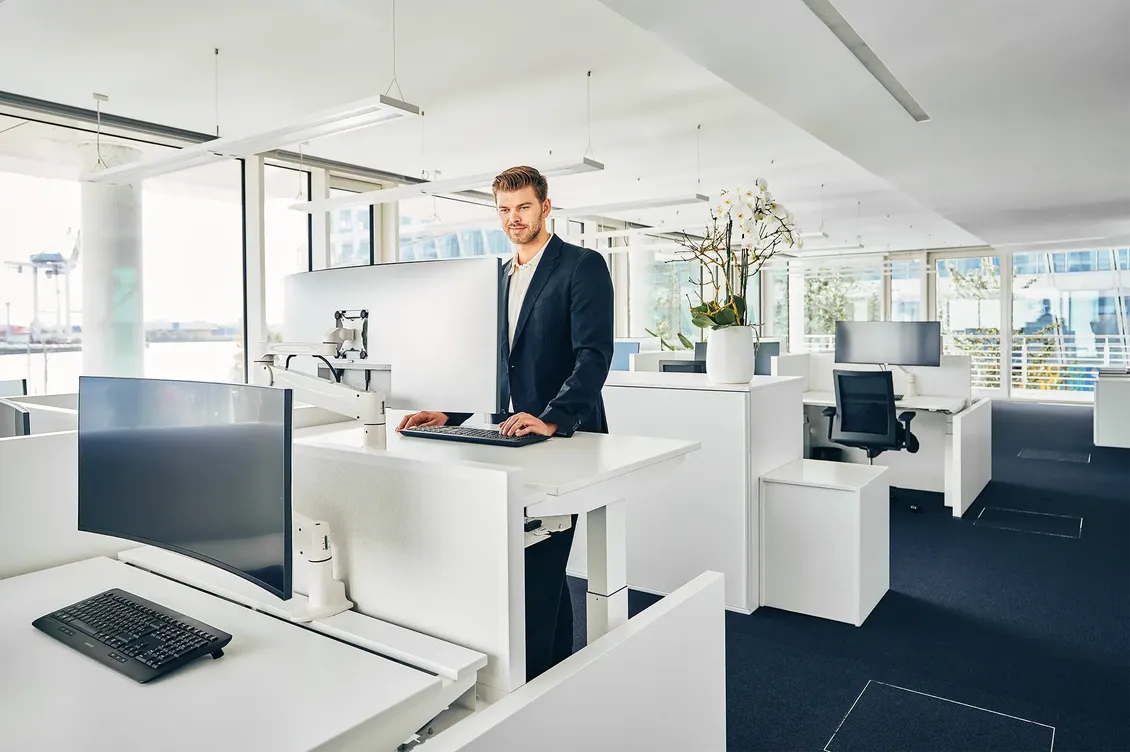 Man in a suit stands at a modern office desk with a computer, surrounded by bright, spacious workstations and large windows.