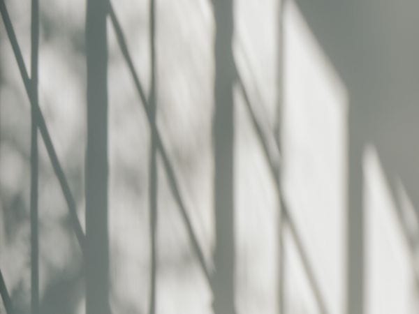 Moderner, minimalistischer Wohnraum mit einer hellen Couch und einem runden Holz-Couchtisch. Durch große Fenster fallen Sonnenstrahlen herein und werfen geometrische Schattenmuster an die graue Wand, was eine ruhige, kontemplative Atmosphäre schafft. Die reduzierte Farbpalette in Grau- und Naturtönen unterstreicht den zeitgenössischen, schlichten Designansatz.